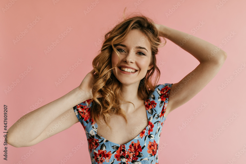 Portrait of blond curly haired woman in blue floral top looking into camera and smiling on isolated backdrop. Modern girl in summer t-shirt posing.. Portrait of blond curly haired woman in blue floral top looking into camera and smiling on isolated backdrop. Modern girl in summer t-shirt posing..