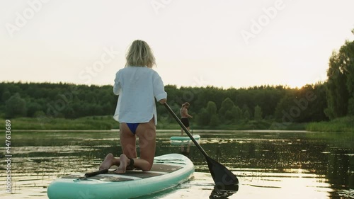 Couple of surfers in lake in sunlught back view, woman beginner paddling after man trainer on surfboards. Sports young family swimming on paddleboards at sunrise. Summer sport, active outdoor vacation photo