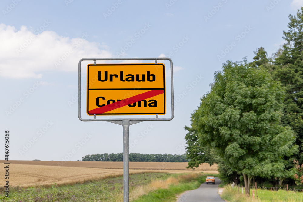Yellow street sign or place name sign with work and vacation in German language. Work is crossed out and in the background you can see a car driving. 