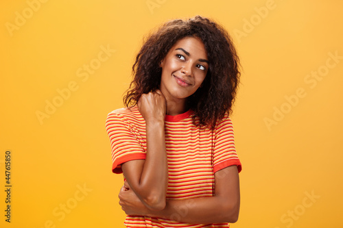 Cute and gentle dreamy dark-skinned girl with cruly hairstyle tilting head smiling from delight and happiness touching neck tender gazing at upper left corner thinking or being nostalgic photo