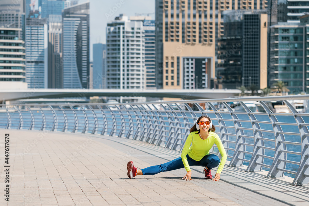 Happy woman wearing headphones stretches and warms up after an intense running workout on the embankment in Dubai Marina