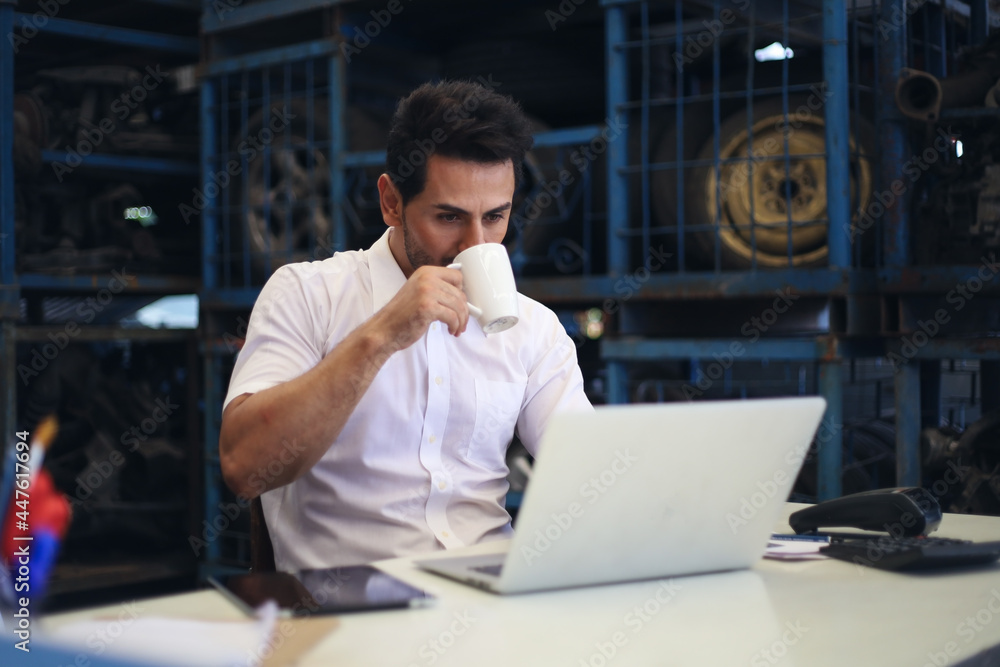 Businessman using laptop computer working or checking stock in warehouse factory, entrepreneurship or business owner relaxing and drinking coffee concept