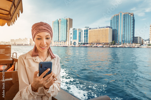 Happy asian woman in a maroon turban using app on her smartphone while cruising on a traditional Abra Dhow boat on Dubai Creek. photo