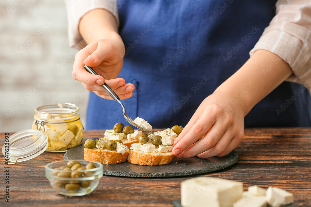 Woman making sandwiches with tasty feta cheese and olives on wooden table in kitchen, closeup