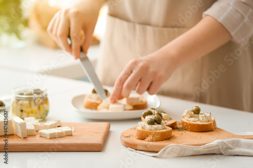 Woman making sandwiches with tasty feta cheese, oil and olives on light table in kitchen, closeup
