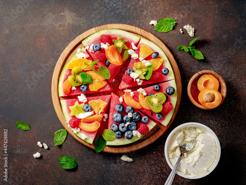 Watermelon pizza bar on dark brown table. Watermelon with assorti of berries, fruits and ricotta cheese. Pieces of watermelon with kiwi, blueberries, raspberries, apricots and orange on wooden board.