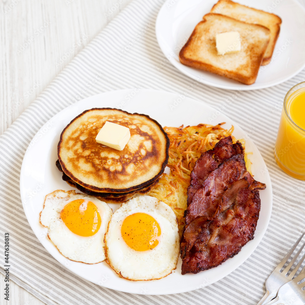 Full American Breakfast with Bacon, Hash Browns, Eggs and Pancakes on a plate on a white wooden surface, side view. Close-up. Full American Breakfast with Bacon, Hash Browns, Eggs and Pancakes on a plate on a white wooden surface, side view. Close-up.