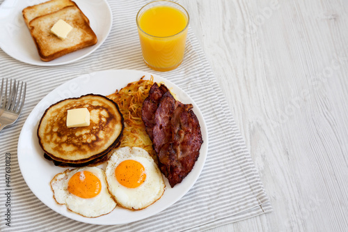 Full American Breakfast with Bacon, Hash Browns, Eggs and Pancakes on a plate on a white wooden table, side view. Copy space.