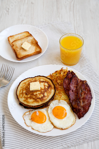 Full American Breakfast with Bacon, Hash Browns, Eggs and Pancakes on a plate on a white wooden background, side view.