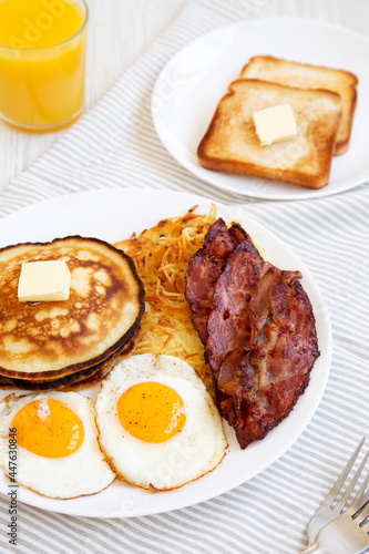 Full American Breakfast with Bacon, Hash Browns, Eggs and Pancakes on a plate on a white wooden table, side view.