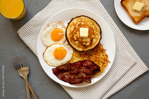 Full American Breakfast with Bacon, Hash Browns, Eggs and Pancakes on a plate on a gray surface, top view.