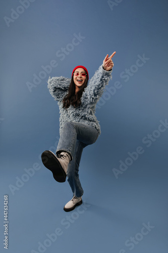 Positive long dark haired lady with red hat and glasses in trendy sweater, blue jeans and light cool boots raising her leg and arm..
