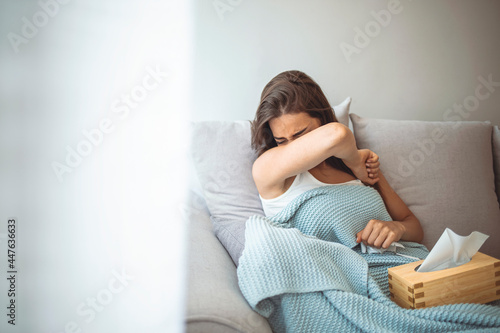 Sick woman sneezing into her elbow while lying down on sofa at home. Coronavirus disease (COVID-19) symptoms - runny nose, sore throat, cough, fever. Woman lying in bed at home quarantine isolation