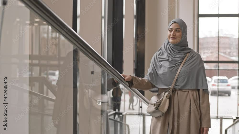 Medium slowmo shot of smiling Arabic woman in hijab looking around while going up escalator in contemporary office building Medium slowmo shot of smiling Arabic woman in hijab looking around while going up escalator in contemporary office building