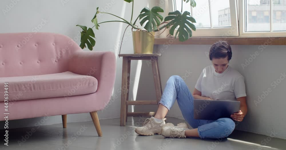 Stylish girl in jeans sitting indoors working on laptop on floor copy space text. Young woman model holding computer notebook at home slow motion. Casual clothes fashion. Self-isolation conditions Stylish girl in jeans sitting indoors working on laptop on floor copy space text. Young woman model holding computer notebook at home slow motion. Casual clothes fashion. Self-isolation conditions