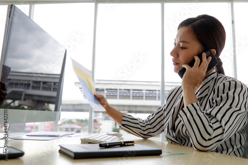 Photography An asian beautiful business woman wearing casual striped pattern shirt, smiling with happiness and talking on mobile or smart phone while looking at computer and sitting in the office