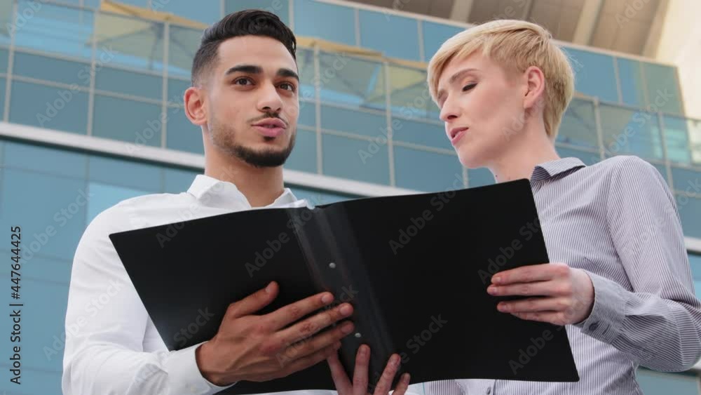 Bottom view arabic man boss leader and caucasian mature adult business woman stand outdoors near company office building holding folder with documents papers contract report discussing talking project Bottom view arabic man boss leader and caucasian mature adult business woman stand outdoors near company office building holding folder with documents papers contract report discussing talking project
