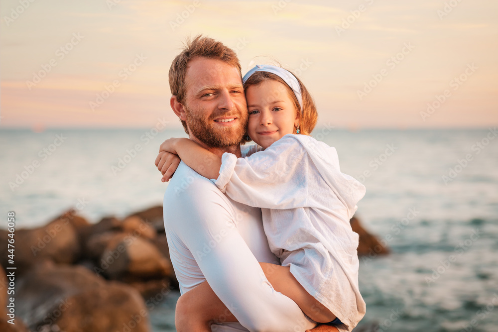 Father's Day. Portrait of a smiling father hugging his preschool-age daughter in his arms. In the background, the sea and the sky. The concept of happy fatherhood and adoption of children Father's Day. Portrait of a smiling father hugging his preschool-age daughter in his arms. In the background, the sea and the sky. The concept of happy fatherhood and adoption of children