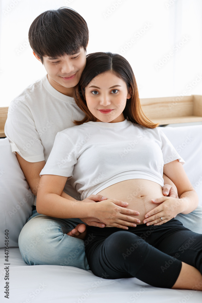 portrait of a lovely young couple dressed casually sitting on a bed together. A beautiful pregnant woman sitting and leaning on a handsome husband's chest happily. Family concept portrait of a lovely young couple dressed casually sitting on a bed together. A beautiful pregnant woman sitting and leaning on a handsome husband's chest happily. Family concept