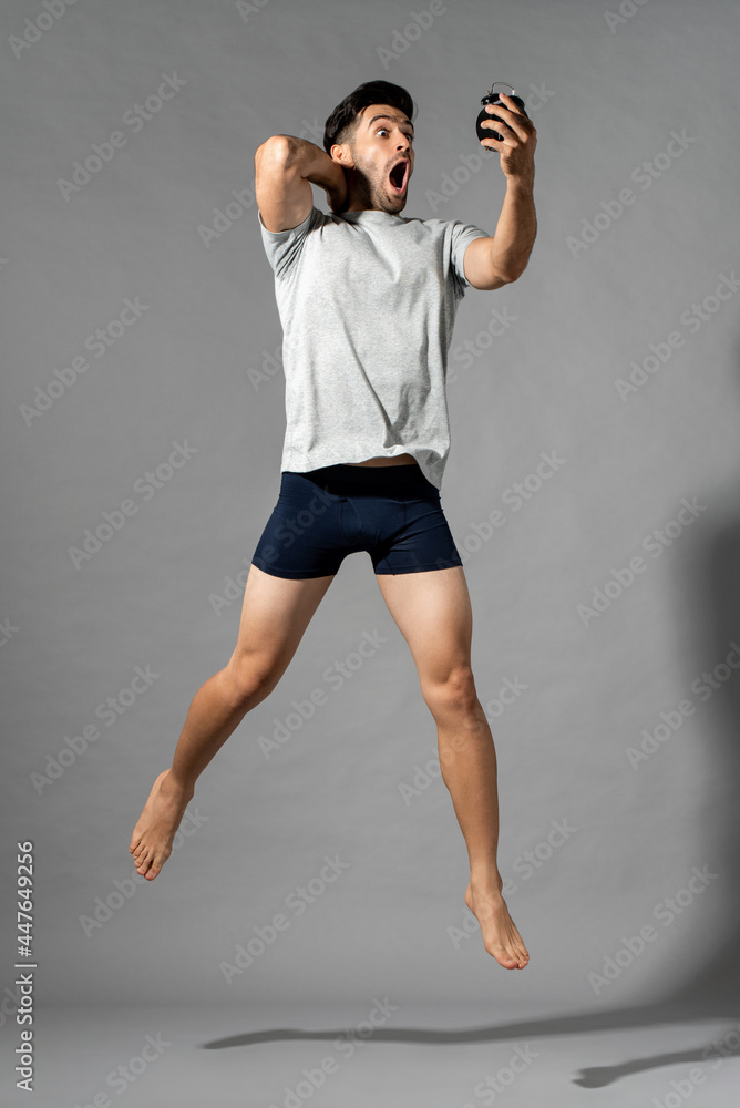 Full length portrait of shocked young Caucasian man in sleepwear waking up late looking at alarm clock and jumping in isolated gray studio background