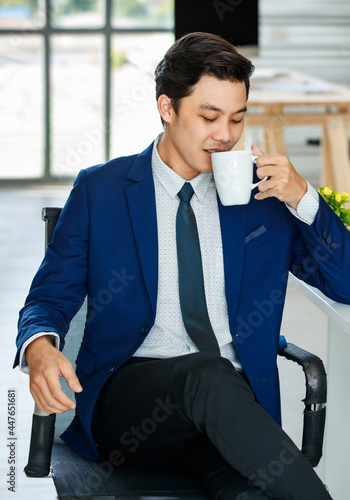 Asian youn businesman in blue suit sitting in office and holding coffee cup with easy and relax pose