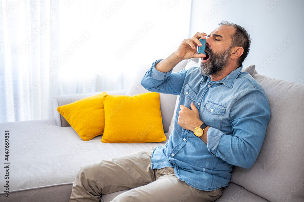 Obraz premium Side view of an asthmatic man using an inhaler sitting on a couch in the living room at home. Man using a pressurized cartridge inhaler extended pharynx, Bronchodilator