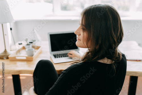 A young freelance businesswoman communicates online works using wireless technologies phone and laptop makes notes in a notebook sitting at a desk in a home office indoors
