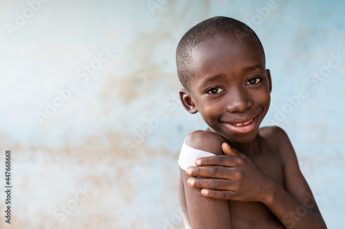 Portrait of a smiling and confident looking bare chested skinny little African boy with a large patch on his shoulder after being vaccinated against childhood infectious disease photo