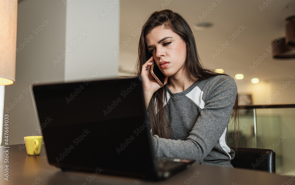 Young beautiful Brazilian woman using smartphone and working with laptop while sitting at office desk, working from home concept. Young beautiful Brazilian woman using smartphone and working with laptop while sitting at office desk, working from home concept.