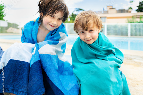 Two brothers are sitting near a pool wrapped in swimming  towels after enjoying water. Children spend summer holidays together in family.