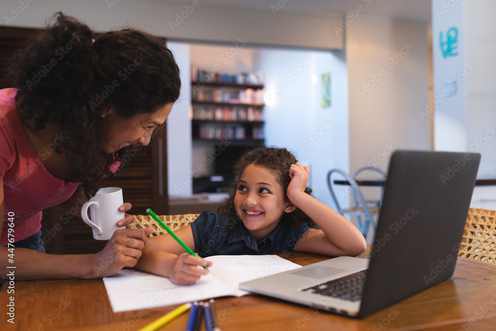 Smiling mixed race mother holding coffee, helping her daughter do homework Smiling mixed race mother holding coffee, helping her daughter do homework