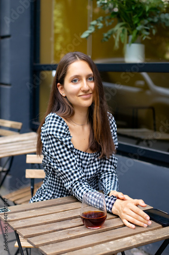fashionable girl in plaid dress looks at the camera and smiles with a drink while sitting at the table in modern outdoor cafe on the background of window