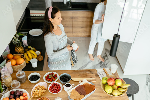 View from above of mature woman with her adult daughter cooking homemade healthy sweets. Different berries and fruits on the table at modern kitchen interior.