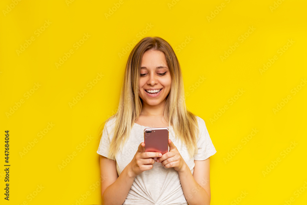 A young caucasian happy pretty blonde woman smiles and holds a mobile phone looking at the screen isolated on a bright color yellow background. The girl uses her smartphone A young caucasian happy pretty blonde woman smiles and holds a mobile phone looking at the screen isolated on a bright color yellow background. The girl uses her smartphone