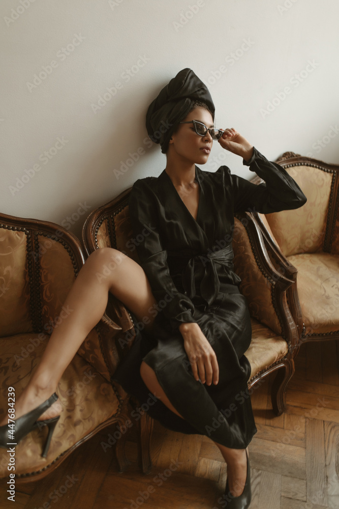 Fototapeta premium Fashionable lady in long sleeve dress and sunglasses posing inside. Modern woman in heels and hat sitting on armchair in apartment..