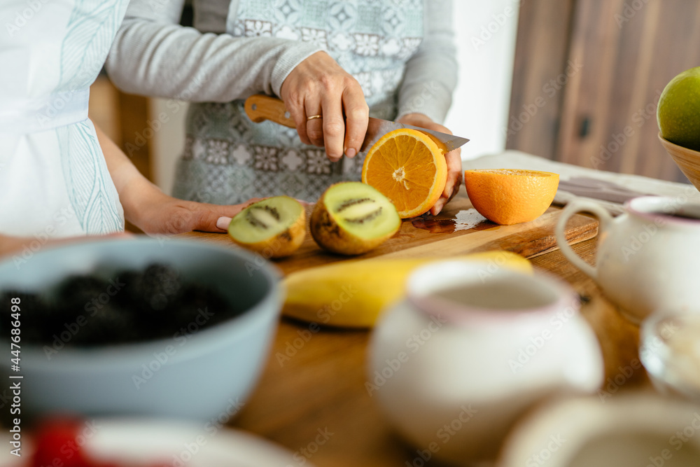 Close up of women hands cooking at home, cutting orange fruit with knife with nuts and seeds, berries and fruits on wooden working surface at kitchen.