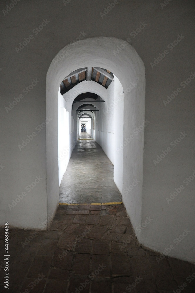An opening and a long corridor in a white wall. Sunlight on a white wall. People in the back of the corridor. 