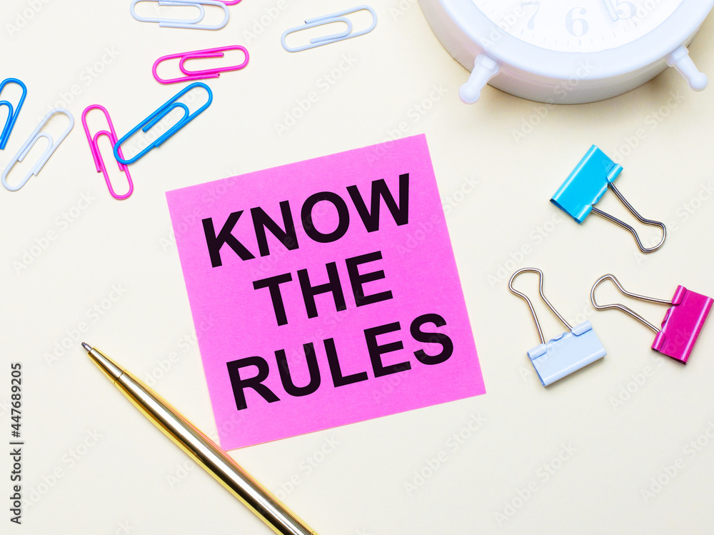 On a light background, a white alarm clock, pink, blue and white paper clips, a golden pen and a pink sticker with the text KNOW THE RULES On a light background, a white alarm clock, pink, blue and white paper clips, a golden pen and a pink sticker with the text KNOW THE RULES