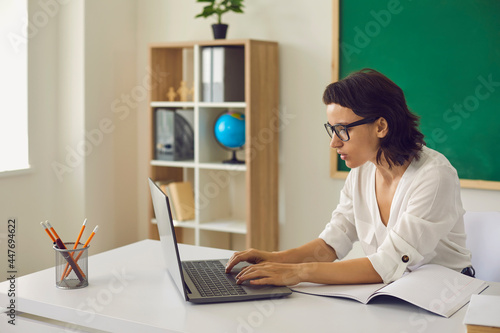 Young brunette woman teacher sitting in classroom and working online on laptop over school classroom furniture background, side view. Online working and education concept photo