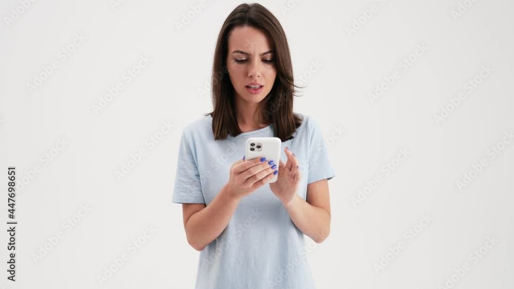 A surprised woman wearing basic t-shirt is using her mobile standing in the studio isolated over white background A surprised woman wearing basic t-shirt is using her mobile standing in the studio isolated over white background