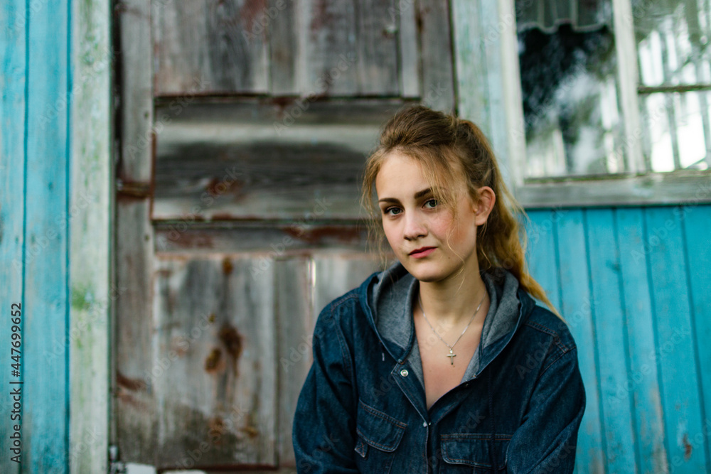 portrait of a young white girl in a denim suit in front of a blue wooden wall of an old village house at sunset