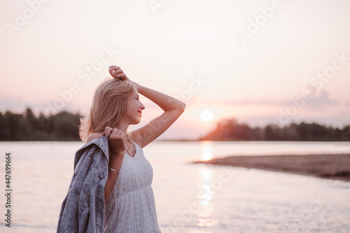 Portrait in profile of a young woman on the beach. A beautiful blonde in a white summer dress stands on the river bank and holds a denim jacket in her hand against the background of sunset and water.