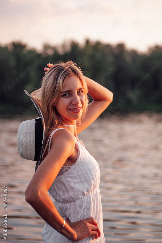 Portrait of a blonde on the background of a river. A beautiful young happy woman poses at sunset in a white dress and a straw hat with a dark ribbon against the background of trees. Portrait of a blonde on the background of a river. A beautiful young happy woman poses at sunset in a white dress and a straw hat with a dark ribbon against the background of trees.