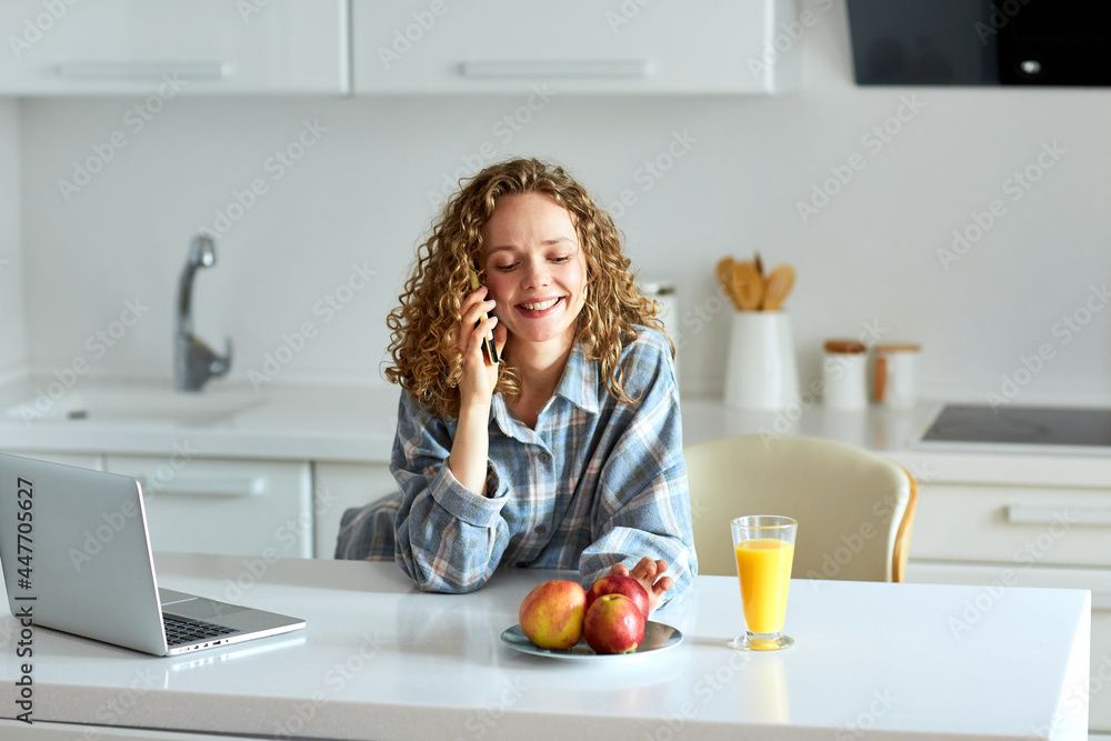 Enjoying time at home. Beautiful young smiling woman working on laptop and drinking orange juice while sitting on the chair in the kitchen at home.Young woman working at home in the morning