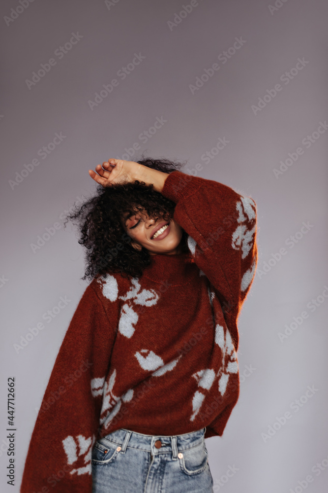 Elegant dark-skinned brunette woman smiles with closed eyes on isolated. Charming curly girl in burgundy sweater poses on grey background. Elegant dark-skinned brunette woman smiles with closed eyes on isolated. Charming curly girl in burgundy sweater poses on grey background.