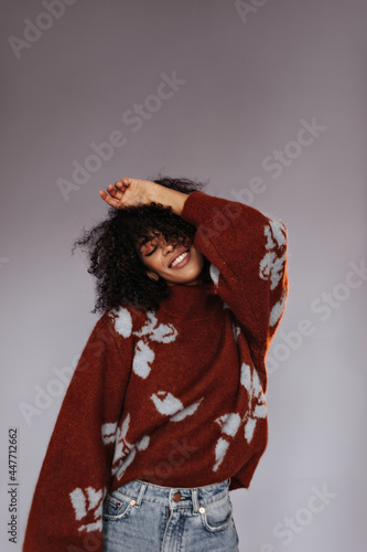 Elegant dark-skinned brunette woman smiles with closed eyes on isolated. Charming curly girl in burgundy sweater poses on grey background.