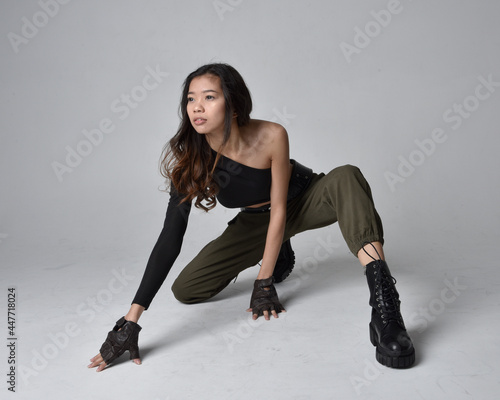Full length portrait of pretty brunette, asian girl wearing black top and khaki utilitarian army pants and leather boots. Sitting pose , isolated agent a light grey studio background.