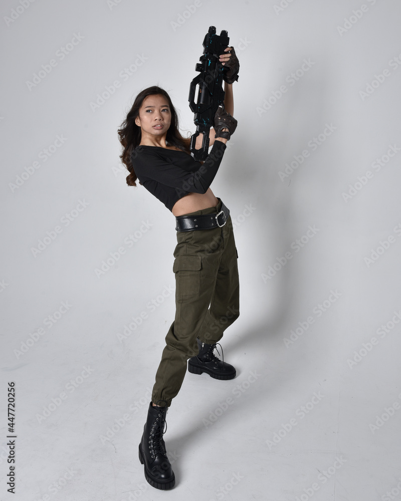 Full length portrait of pretty brunette, asian girl wearing black top and khaki utilitarian army pants and leather boots. Standing pose holding a science fiction gun, isolated agent a light grey studi Full length portrait of pretty brunette, asian girl wearing black top and khaki utilitarian army pants and leather boots. Standing pose holding a science fiction gun, isolated agent a light grey studi