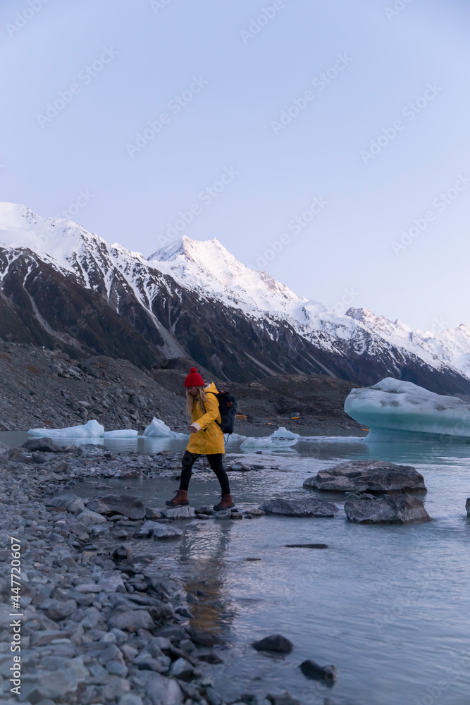 Travel lifestyle view of girl hiking at foot of alpine Tasman Lake with icebergs at sunrise and Mount Cook mountain, in Mount Cook National Park, South Island of New Zealand.