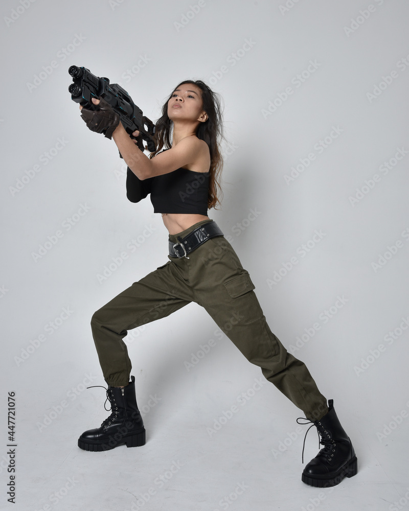 Full length portrait of pretty brunette, asian girl wearing black top and khaki utilitarian army pants and leather boots. Standing pose holding a science fiction gun, isolated agent a light grey studi Full length portrait of pretty brunette, asian girl wearing black top and khaki utilitarian army pants and leather boots. Standing pose holding a science fiction gun, isolated agent a light grey studi